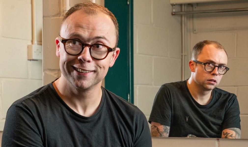 Joe Tracini, presenter of the documentary, smiling in the foreground, looking sad and worried in the background mirror